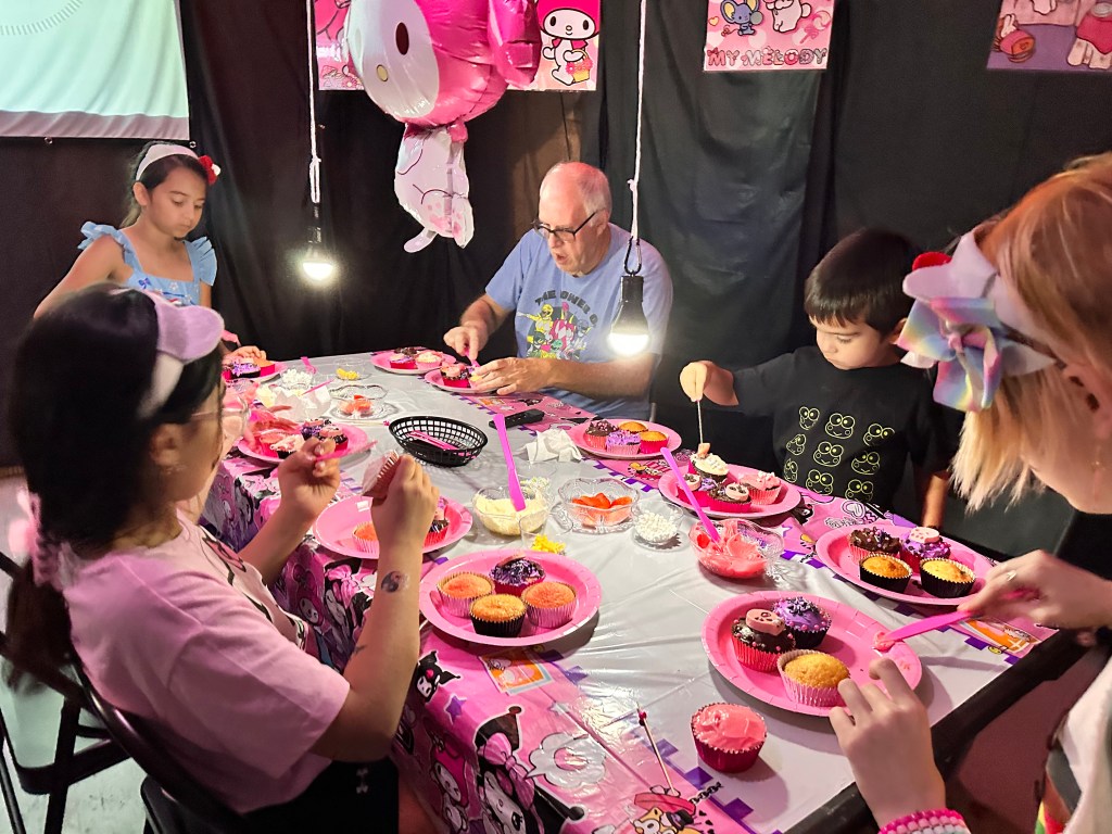 A group of kids, plus an adult honorary kid, decorating cupcakes during a Hello Kitty cupcake wars party activity, sitting at a Kuromi and My Melody themed table.