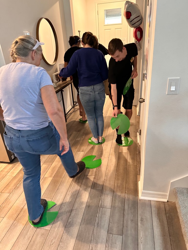 Adults balancing on green lily pads cut from card stock as they race through a hallway during a Sanrio-themed party game.