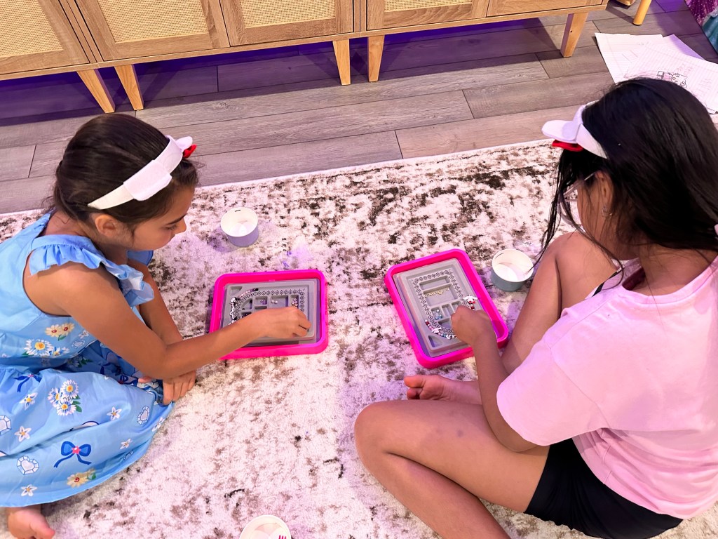 Two girls sitting on the floor with pink trays, carefully assembling their Sanrio-themed beaded bracelets.