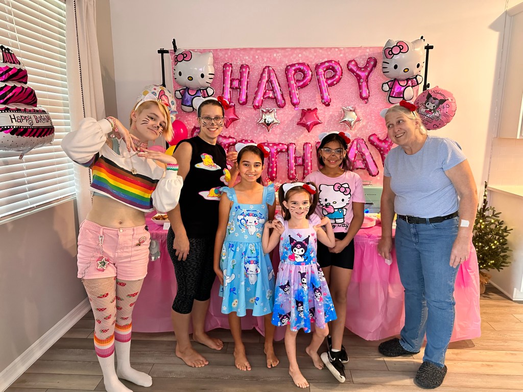 A group of party guests smiling in front of a pink “Happy Birthday” backdrop, with several wearing Hello Kitty ears and whiskers painted on their faces.