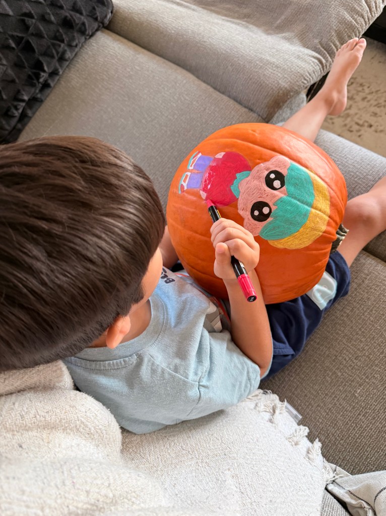 A young boy colors a pumpkin with a chibi Baby Saja character and a soda pop can using paint markers during a K-Pop Demon Hunters themed family craft activity.