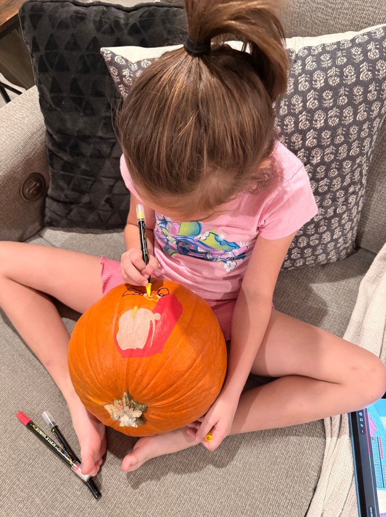 A young girl in a pink shirt carefully paints Mira from K Pop Demon Hunters onto a Halloween pumpkin using acrylic markers.