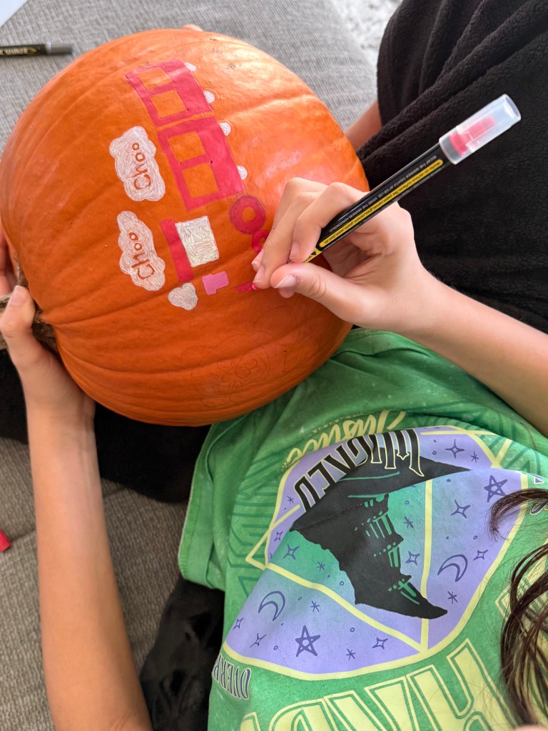 A child uses acrylic paint markers to color a pumpkin featuring a red train and “Choo Choo” clouds, part of a K-Pop Demon Hunters themed craft.