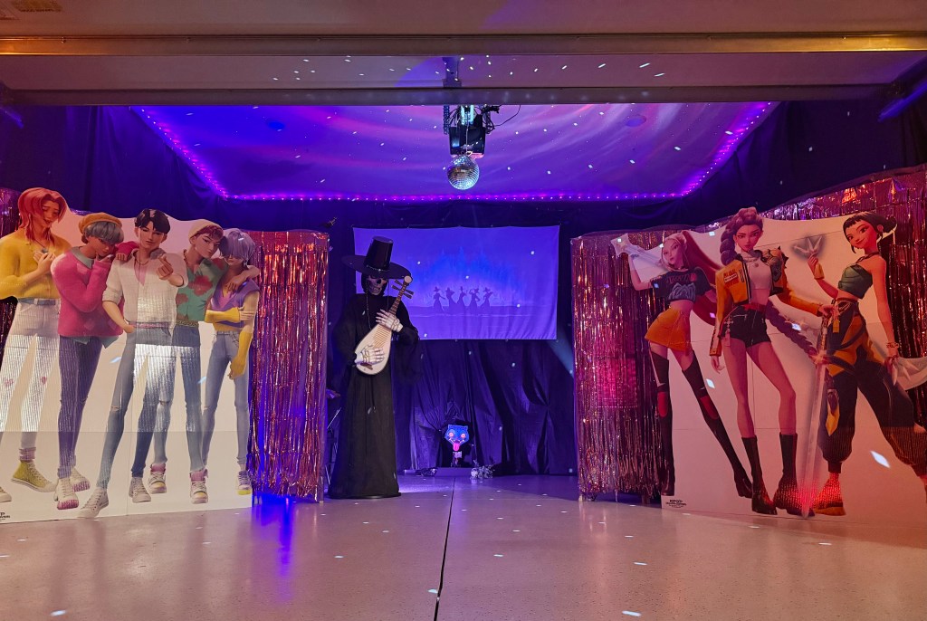 Transformed Halloween garage decorated as a K-Pop Demon Hunters headquarters, featuring life-size cardboard standees of animated K-pop characters, a skeleton musician with a Korean-style bipa, purple LED lighting, disco ball reflections, and a glowing projector screen with shadowed dancers in the background.