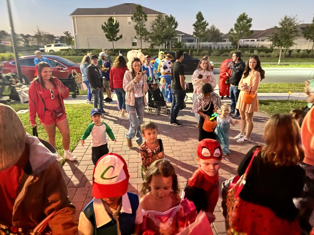 A crowd of costumed children and parents fills the driveway on Halloween night, eagerly approaching the K-Pop Demon Hunters themed garage setup, with families smiling, taking photos, and trick-or-treating in a lively neighborhood scene.