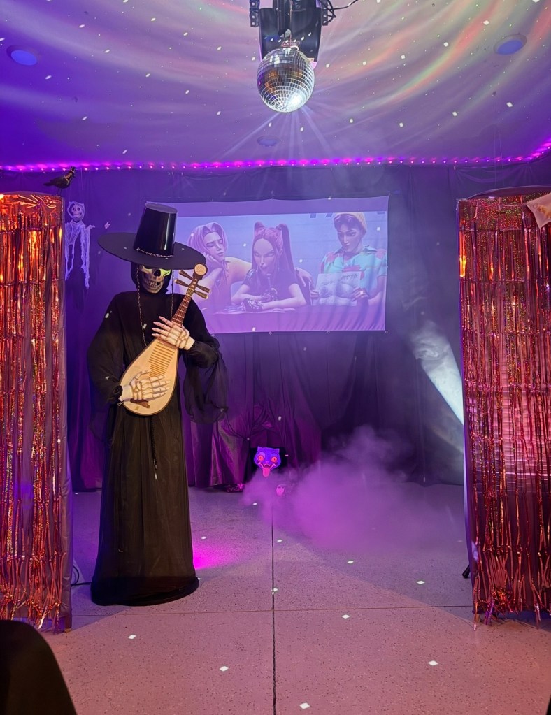 A fully immersive K-Pop Demon Hunters Halloween garage setup featuring a skeleton version of Jinu holding a bipa, dramatic fog effects, a disco ball casting reflections, and a movie projection screen framed by shimmering metallic curtains.