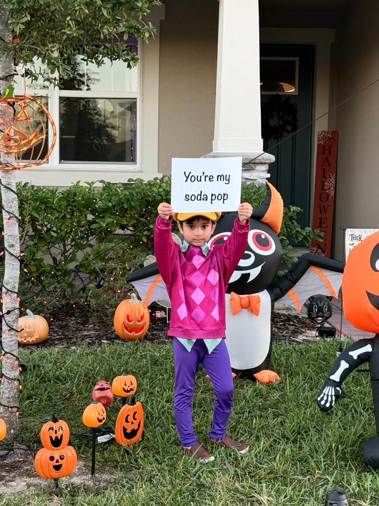 A young boy dressed as Baby Saja from K-Pop Demon Hunters holds up a “You’re my soda pop” quote sign in the front yard beside Halloween inflatables and jack-o’-lanterns.