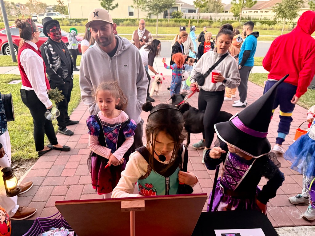 A group of trick-or-treaters in costume gathers around the K-Pop Demon Hunters personality test game, with children eagerly playing and adults watching during a busy Halloween night event.
