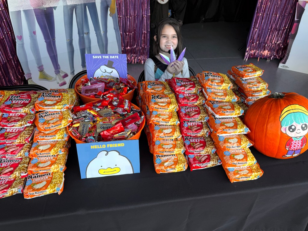 Stacks of Top Ramen packets and two bowls of Halloween candy arranged on a treat table with “Save the Date” and “Hello Friend” themed signs, alongside a painted Baby Saja pumpkin and a smiling child in costume at the K-Pop Demon Hunters Halloween garage.