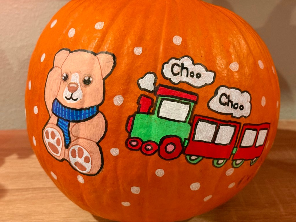 A pumpkin decorated with a cozy teddy bear wearing a scarf next to a red and green train labeled “Choo Choo,” part of a themed painting project.