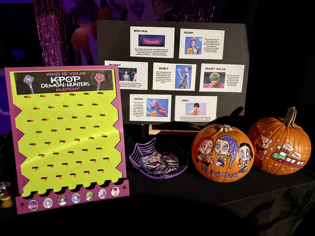 A K-Pop Demon Hunters themed personality match game featuring a colorful disk drop board and a character trait display, set on a black table alongside painted pumpkins and collectible photo cards during Halloween 2025.