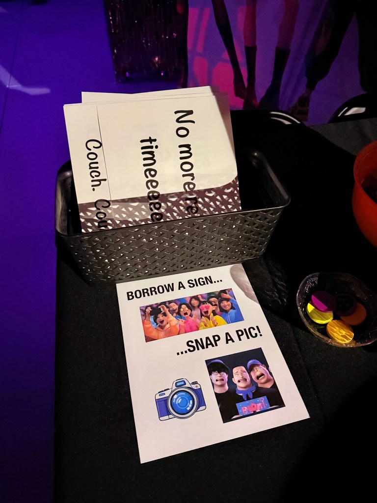 A photo booth station with K-Pop Demon Hunters quote signs in a black basket, a display sign encouraging guests to borrow a sign and take a photo, and colorful game tokens on a Halloween-decorated table.