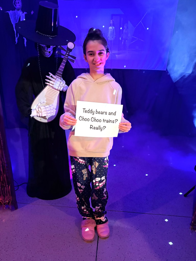 A girl wearing teddy bear pajama pants and a hoodie stands in the Halloween garage holding a K-Pop Demon Hunters quote sign that says “Teddy bears and Choo Choo trains? Really?” with a skeleton Jinu figure and purple lighting in the background.