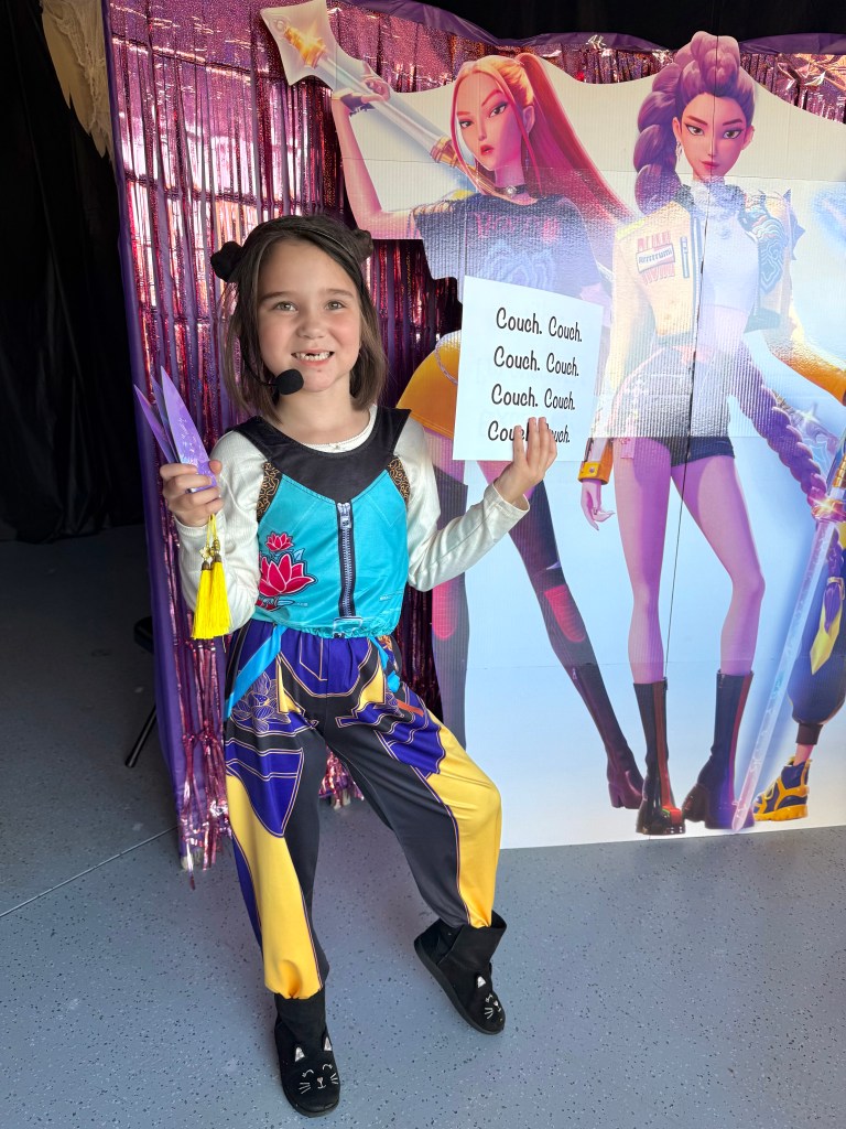 A young girl dressed as Zoey from K-Pop Demon Hunters poses in front of a cardboard backdrop holding a quote sign that reads “Couch. Couch. Couch.” while smiling.
