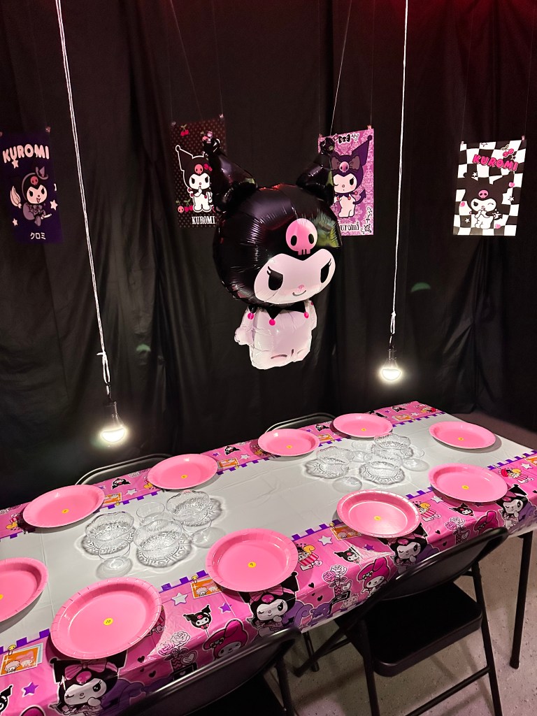 Close view of the Kuromi table with pink plates, clear topping bowls, and a large Kuromi balloon hanging above against black drapes.
