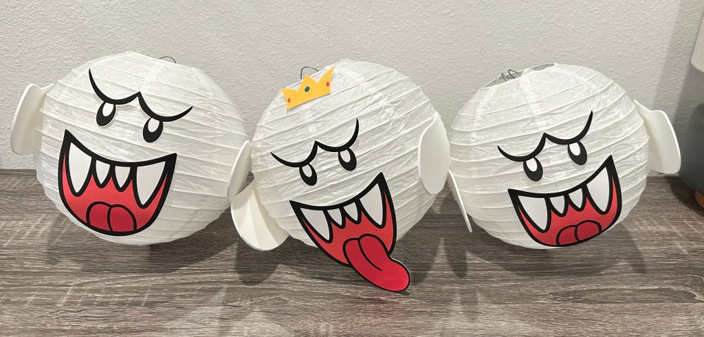 Three Boo ghost decorations made from white paper lanterns, featuring printed face pieces and white craft foam arms, with one ghost wearing a crown to represent King Boo.