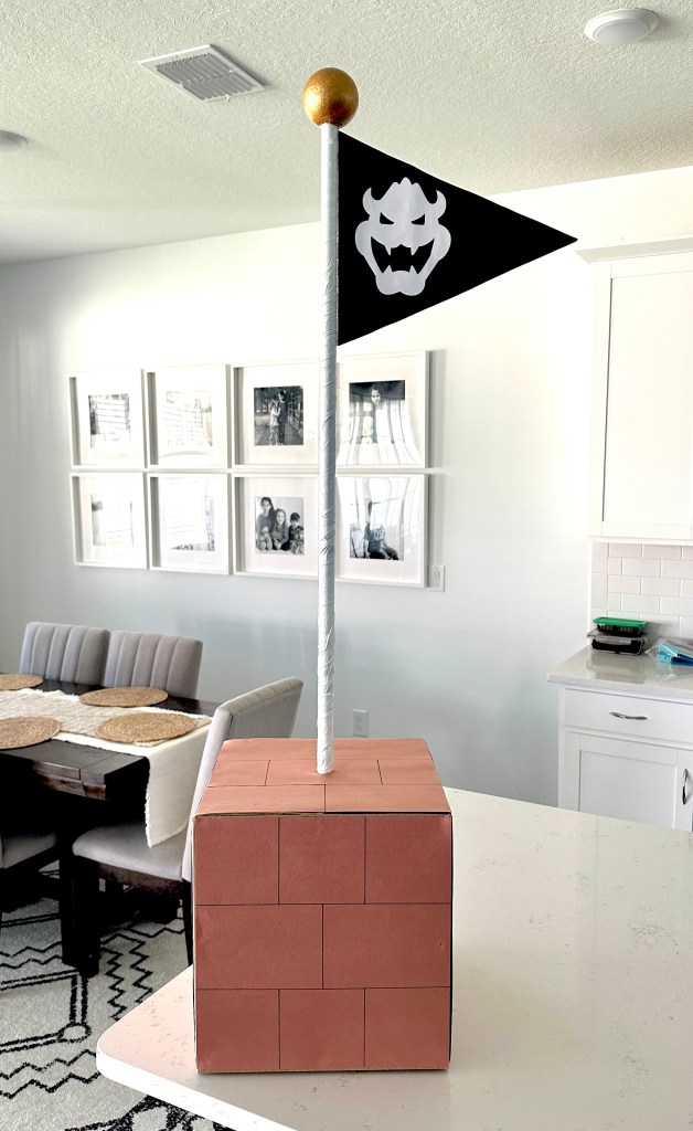 DIY Bowser’s Flag decoration featuring a brick block base, white pole, and black pennant with Bowser emblem for a Mario-themed birthday party