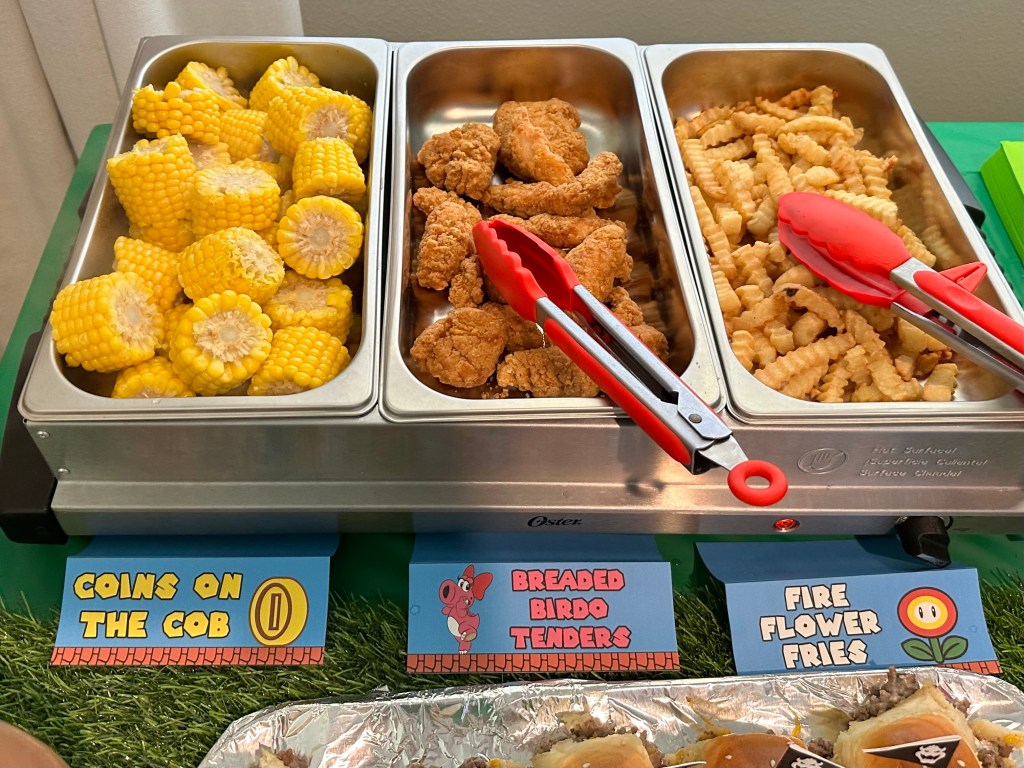 Hot food tray featuring mini corn cobs, chicken tenders, and crinkle fries with themed labels: “Coins on the Cob,” “Breaded Birdo Tenders,” and “Fire Flower Fries.”