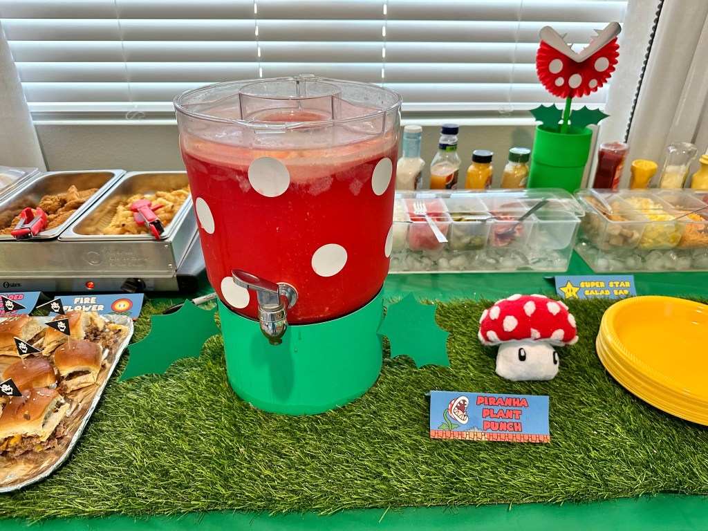 DIY Piranha Plant punch dispenser and matching plush mushroom on a Mario-themed drink station table with food cards and greenery.