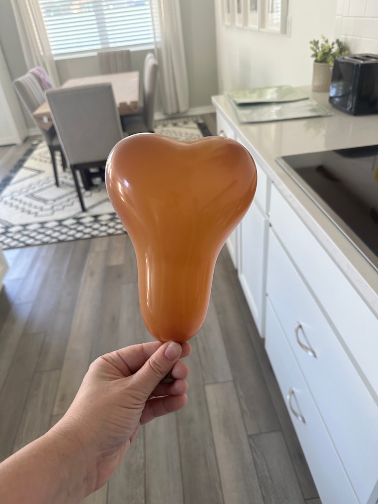 A hand holding an incorrectly inflated mocha brown heart-shaped balloon which created an interesting shape.