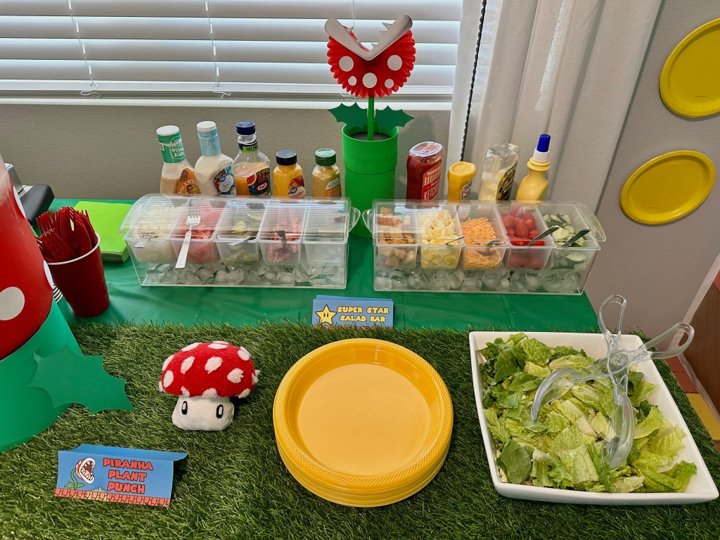 Close-up of Mario-themed salad bar with dressing bottles and chopped toppings on ice, featuring themed food card for “Super Star Salad Bar.”