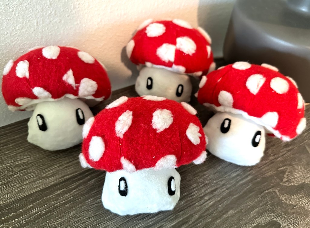 A group of plush red-topped mushrooms customized with small black and white felt eyes to resemble Mario mushrooms, displayed on a wooden surface.