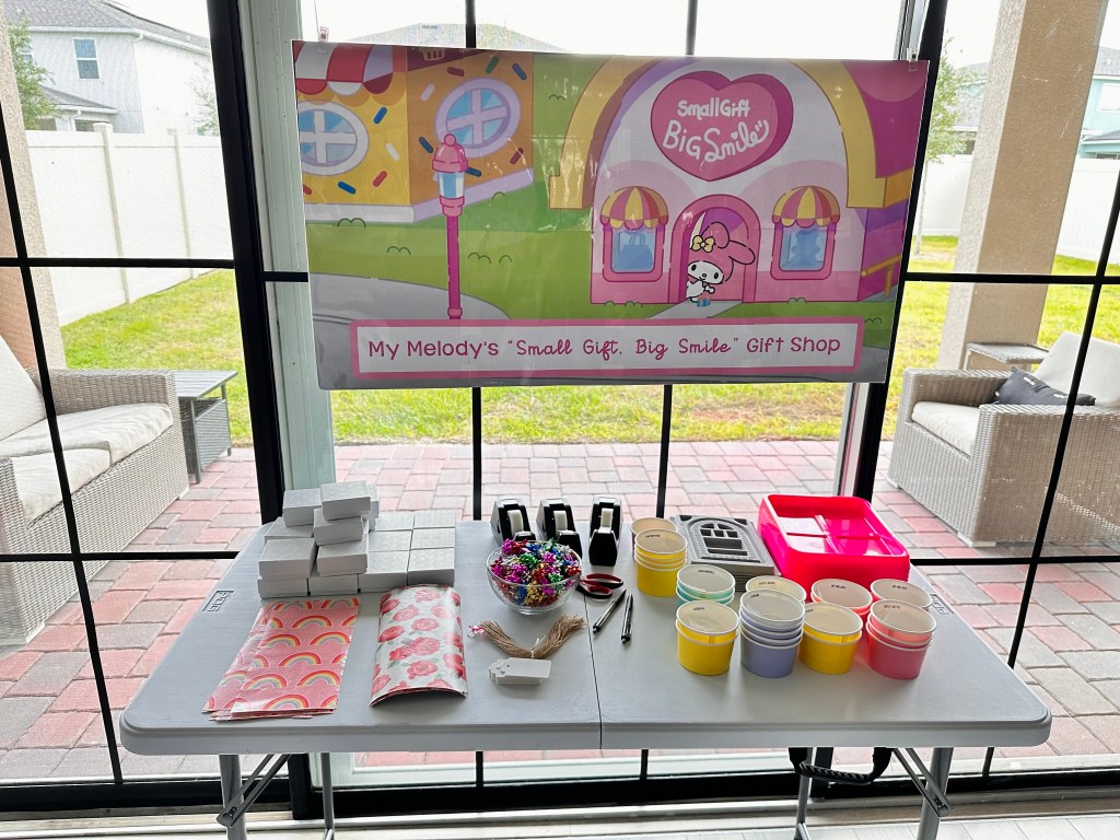 A table display with gift wrap, boxes, ribbon, and a sign reading “My Melody’s Small Gift, Big Smile Gift Shop.”