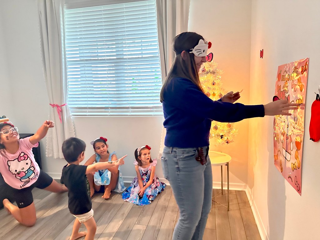A blindfolded young woman reaches toward a Hello Kitty poster while several kids seated nearby point in the wrong direction.