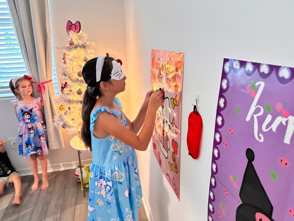 A blindfolded girl places a bow near the correct spot on a Hello Kitty poster while her teammates look on proudly.