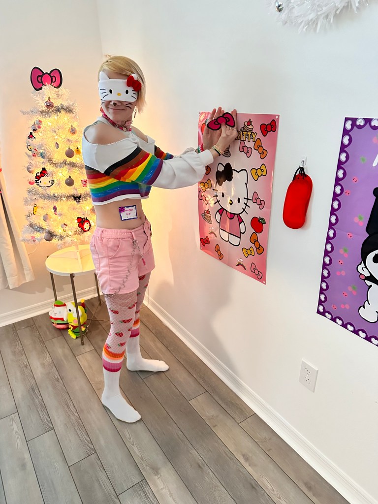 Child wearing a Hello Kitty blindfold attempts to pin a bow onto a Hello Kitty poster, guided by other kids in the background.