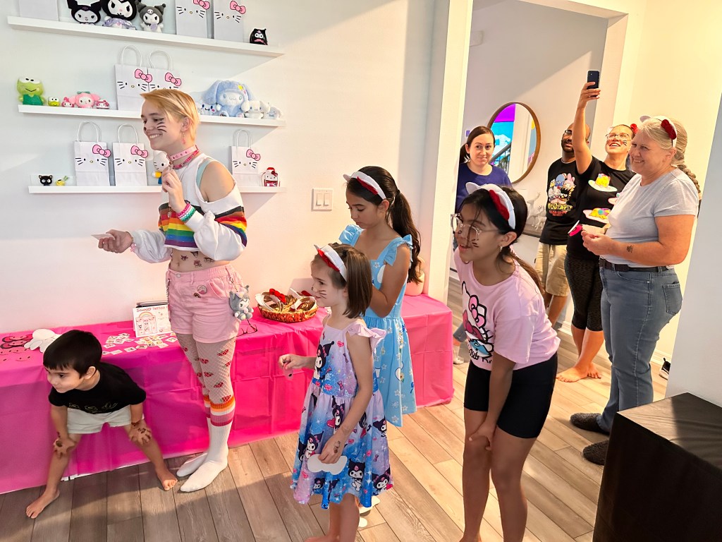 A group of kids and adults watching someone play “Pin the Bow on Hello Kitty,” many smiling or laughing.