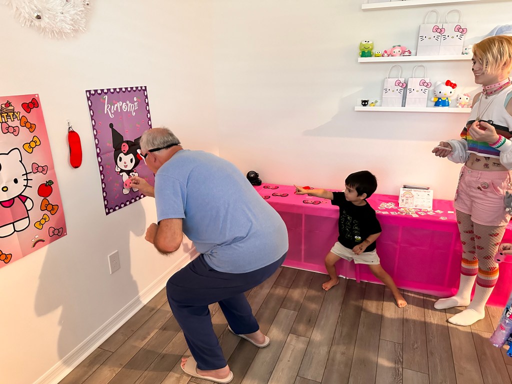 An older man in a blue shirt pins a bow on a Kuromi poster while a small boy points and directs him; a pink table and character decor are in the background.