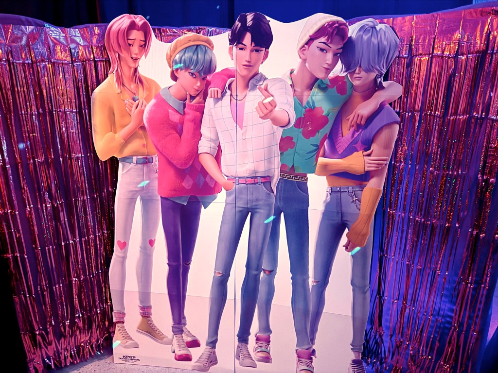Close-up of the Saja Boys standees framed by sparkling pink foil curtains, creating a K-Pop concert vibe inside the Halloween garage.