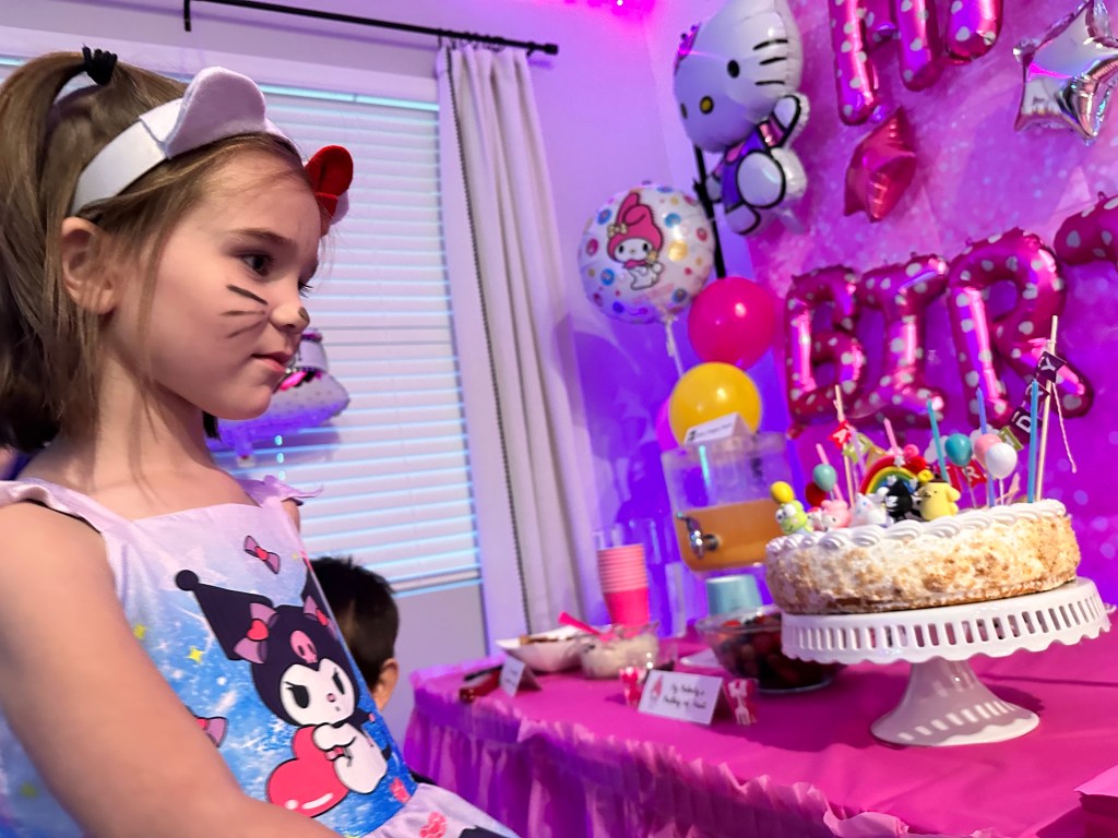 A young girl dressed in a Kuromi-themed dress and cat ear headband smiles while standing near a decorated Sanrio-themed birthday cake topped with colorful character figurines. The cake sits on a white pedestal in front of a pink birthday backdrop with balloons and party decorations.