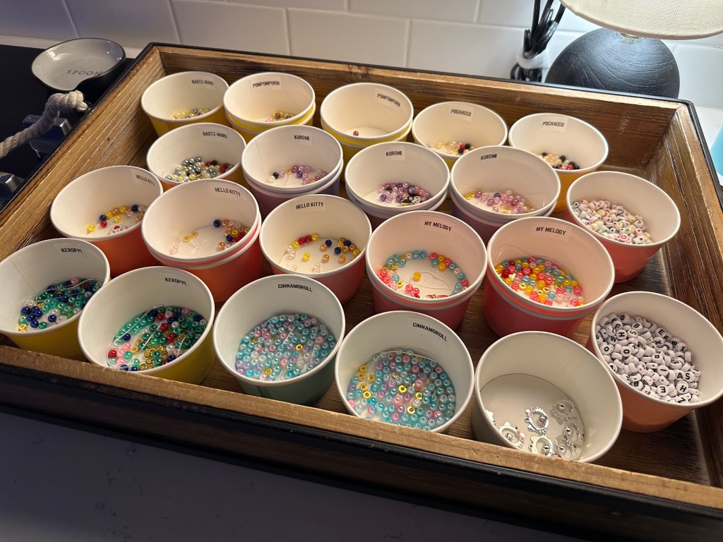 A tray of labeled cups filled with different colored beads, each cup named after a Sanrio character like Hello Kitty, Kuromi, Cinnamoroll, and Keroppi.