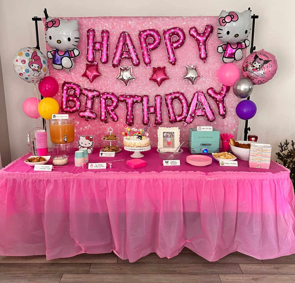 Pink-themed food table with Hello Kitty mylar balloons, a “Happy Birthday” balloon backdrop, and Sanrio-themed food and drink stations.