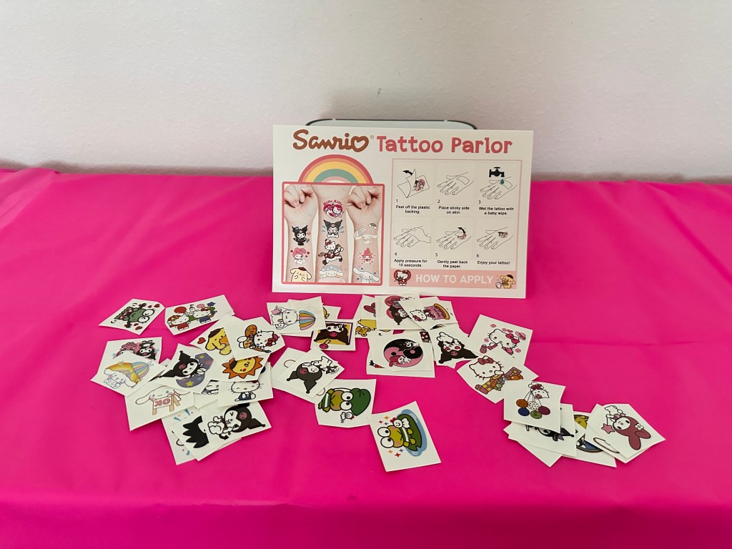 Sanrio Tattoo Parlor setup on a hot pink tablecloth with a printed sign showing application steps and a spread of colorful Sanrio character tattoos.