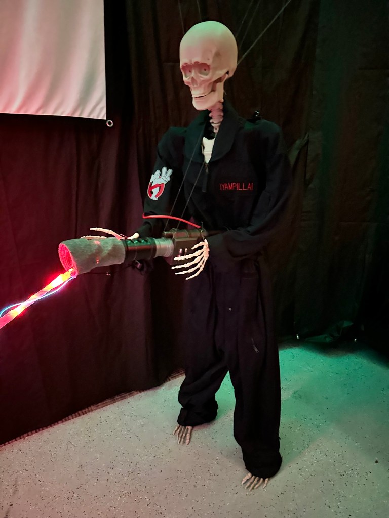  Skeleton dressed in a custom Ghostbusters uniform holding a handmade proton wand with red LED lights in a Halloween garage setup.