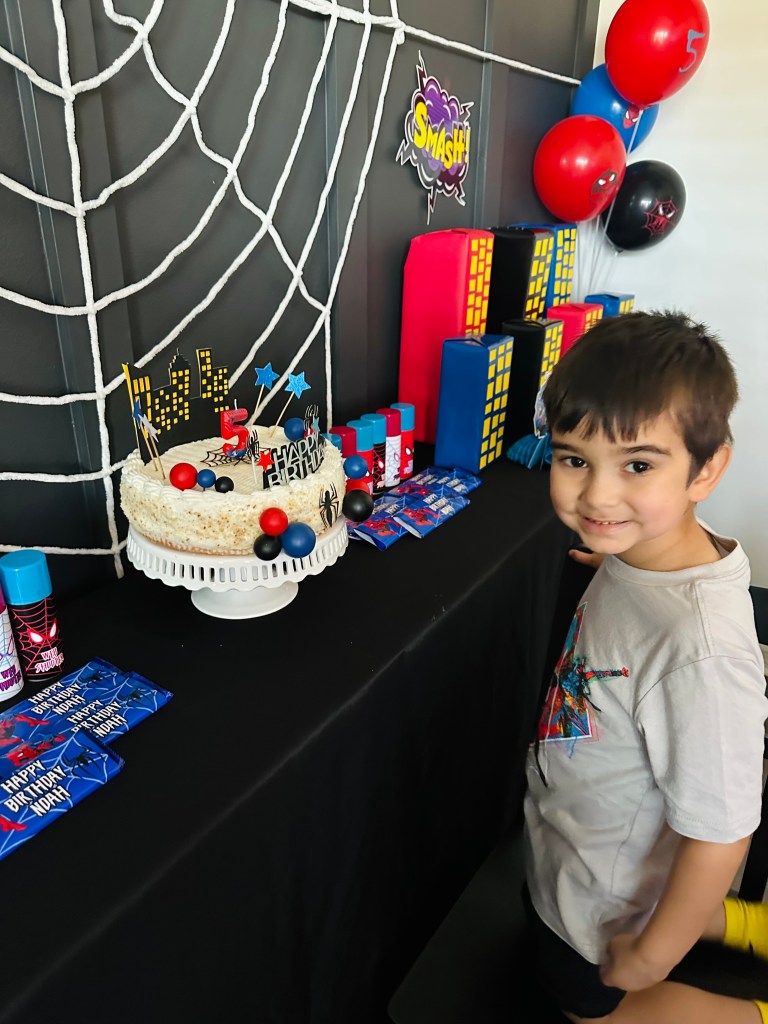 Young boy smiling beside a Spider-Man-themed cake table with DIY city buildings, balloons, web shooters, and a decorated cake centerpiece.