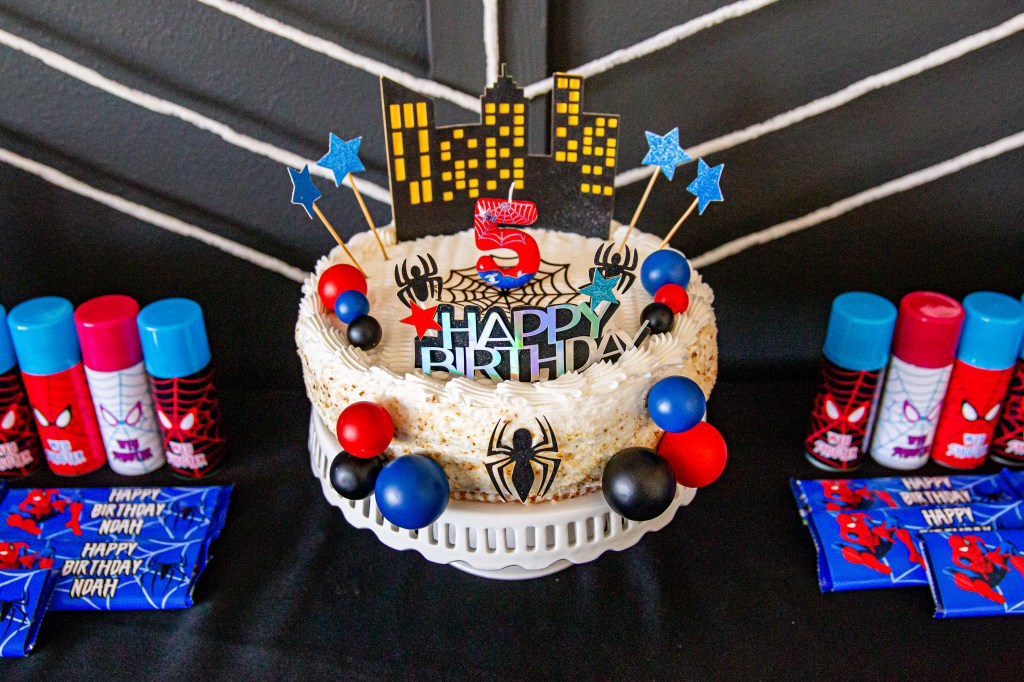Close-up of a Spider-Man birthday cake decorated with cityscape toppers, spider cutouts, red and blue ball accents, and a number 5 candle, surrounded by web shooters and candy bars.