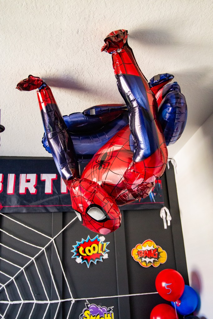 Life-sized Spider-Man balloon hanging upside down from the ceiling, surrounded by comic-style wall decorations and balloons.