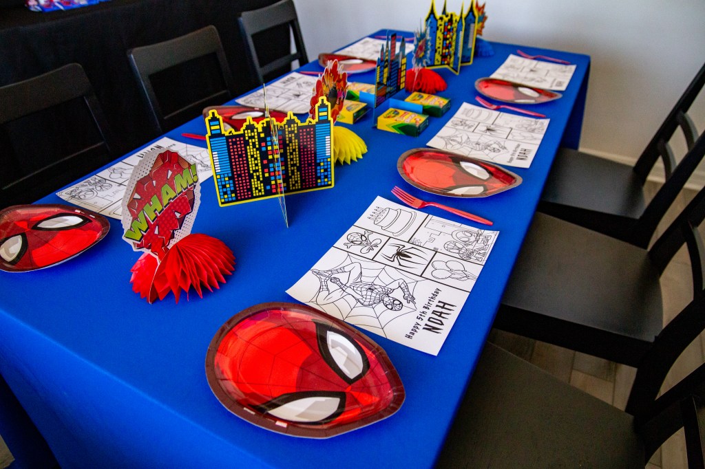 Kids’ party table decorated with red Spider-Man plates, blue tablecloth, cityscape centerpieces, and DIY coloring placemats that say “Happy 5th Birthday Noah.”