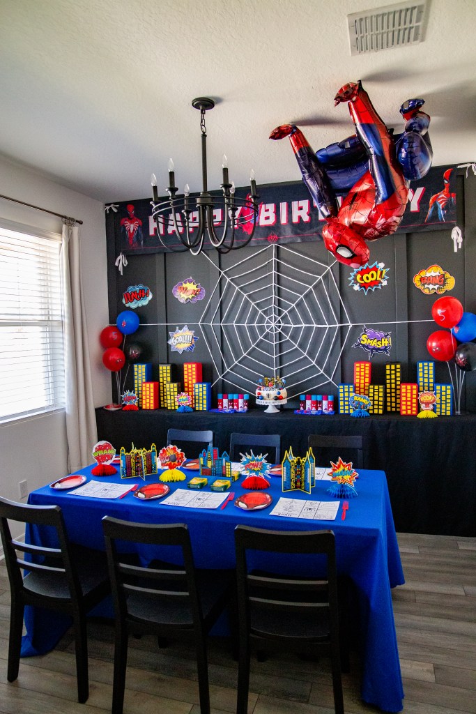 Spider-Man themed party room with comic book city buildings, spider web backdrop, balloons, and a large Spider-Man balloon hanging upside down from the ceiling.