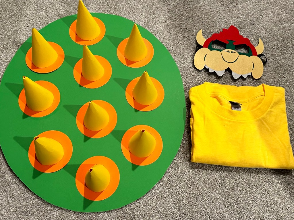 DIY Bowser’s Shield costume piece made from green and orange cardstock, displayed with a Bowser mask and yellow shirt for a Super Mario party.
