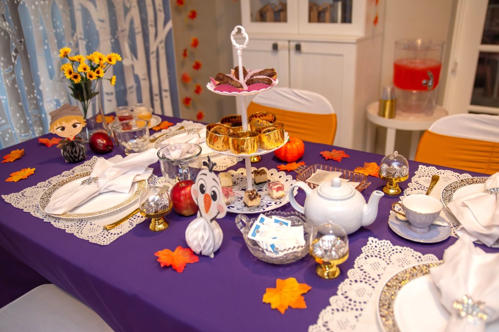 End-of-table Frozen 2 tea party setup with Olaf and Kristoff paper characters, tiered food tray, white teapot, candy domes, apples, and small pumpkins atop a purple tablecloth.