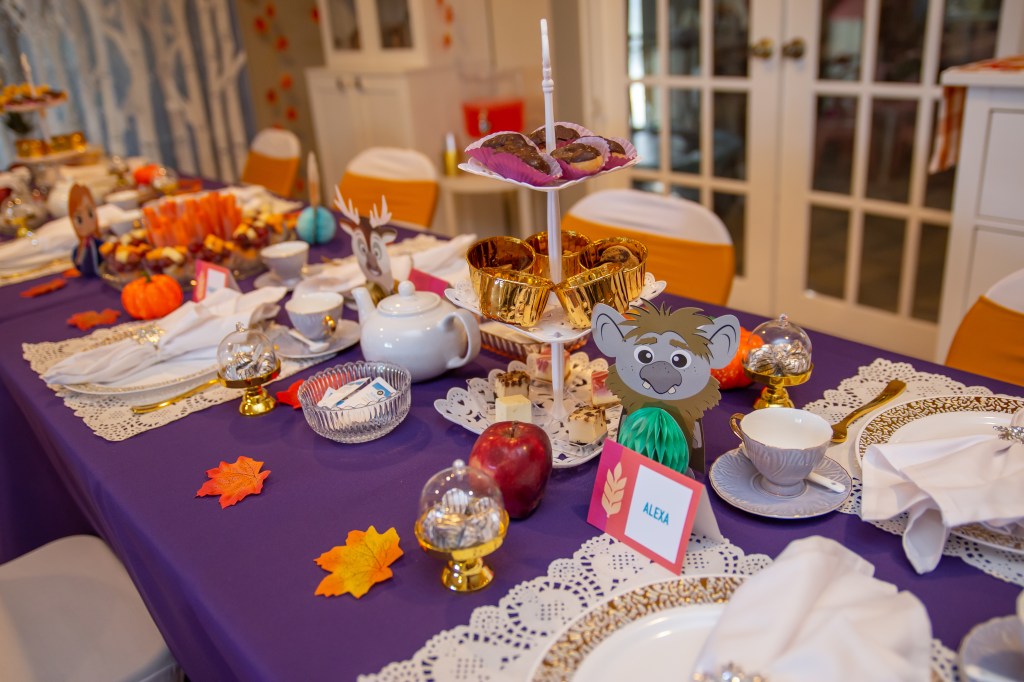 Wide shot of the Frozen 2-themed tea party table decorated with tiered treat stands, small paper Frozen characters, faux apples, mini pumpkins, scattered autumn leaves, and a rich purple tablecloth with lace placemats.