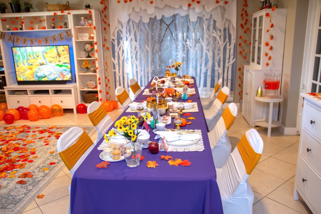 Wide view of a Frozen 2-themed tea party table setup with a birch forest backdrop, autumn leaves, and character-themed seating for children. Purple tablecloth and orange accents evoke a cozy fall atmosphere.