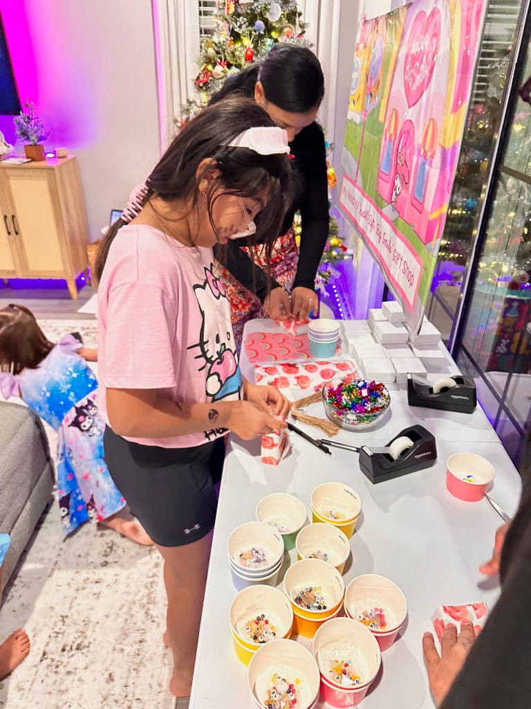 Guests standing at a table wrapping their finished bracelets using colorful gift wrap and tape, with Sanrio beads and cups in the foreground.