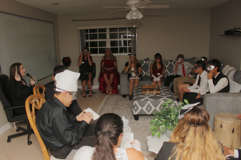 Abby with a microphone, presenting Round 1 of the murder mystery game while guests in formal attire and masks listen in a dimly lit living room with a large screen.
