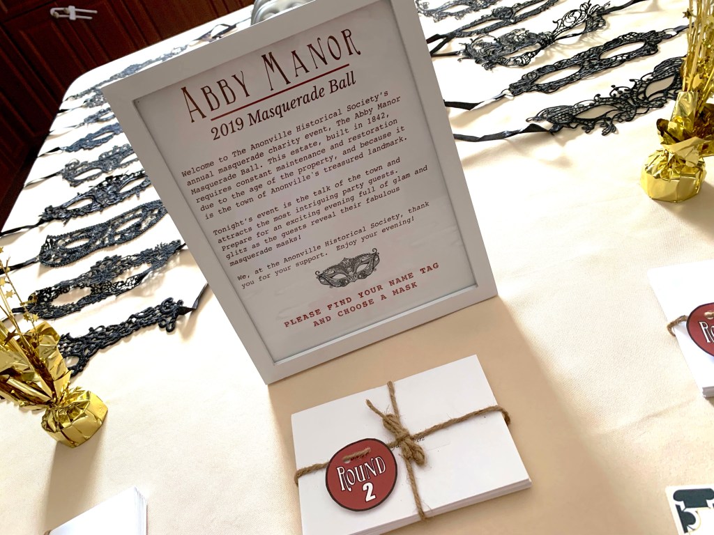 A framed welcome sign for the Abby Manor 2019 Masquerade Ball sits on a table surrounded by masquerade masks and bundles of clue cards labeled “Round 2.”